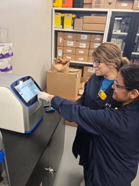 Two scientists, wearing blue lab coats and safety glasses, are standing in a lab. The scientist on the right is pointing to the screen of a modern scientific instrument, and the other scientist is looking at the screen. The lab has shelves in the background, which are filled with various lab supplies.