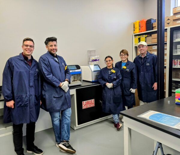 A group of five scientists, all wearing blue lab coats, are standing together in a lab. In the center of the group are two female scientists, and on either side are three male scientists. A large scientific machine is on the counter behind them.