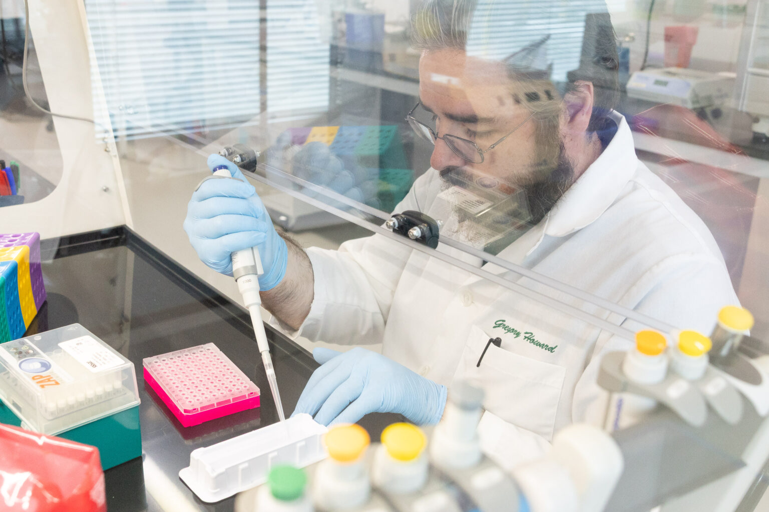 A male scientist wearing a lab coat and blue gloves uses a pipette to transfer a liquid into a tray of small sample wells while working at a laboratory bench.
