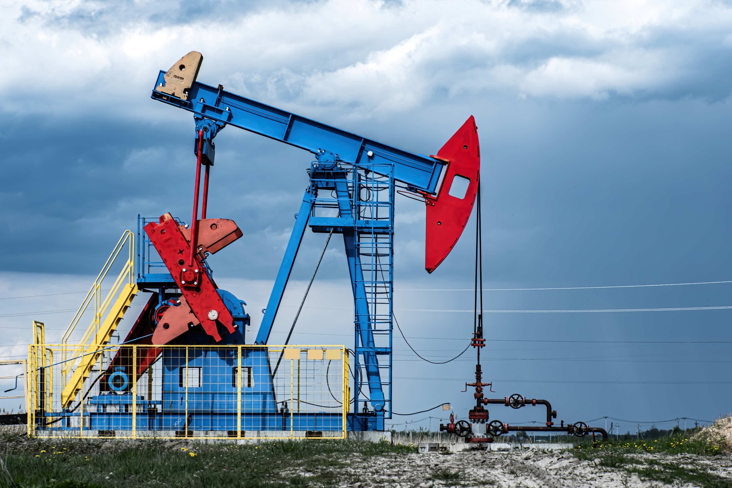 A brightly colored oil pumpjack operating in an open oil field under a cloudy sky. The pumpjack is painted in blue and red with a yellow stairway and safety fence surrounding it.