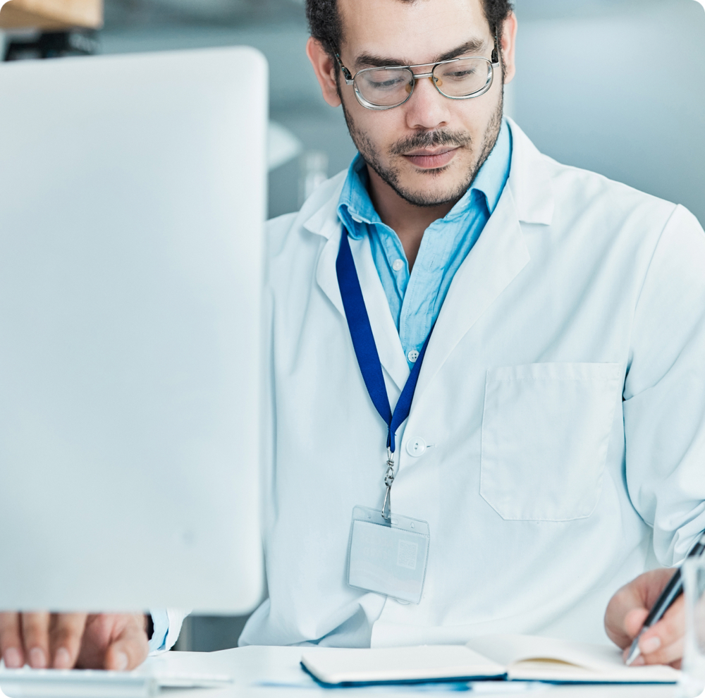 man in lab coat in front of a computer, writing something on a piece of paper